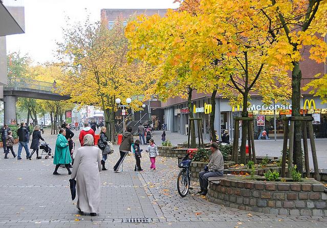 1877_1085 Billstedter Marktplatz im Herbst - Bilder aus den Stadtteilen Hamburgs. Strassenbume stehen in gemauerten Rotunden, am Rand sind Sitzbnke aus Holz eingelassen. Passanten gehen ber den Platz - Kinder spielen unter den Herbstbumen.