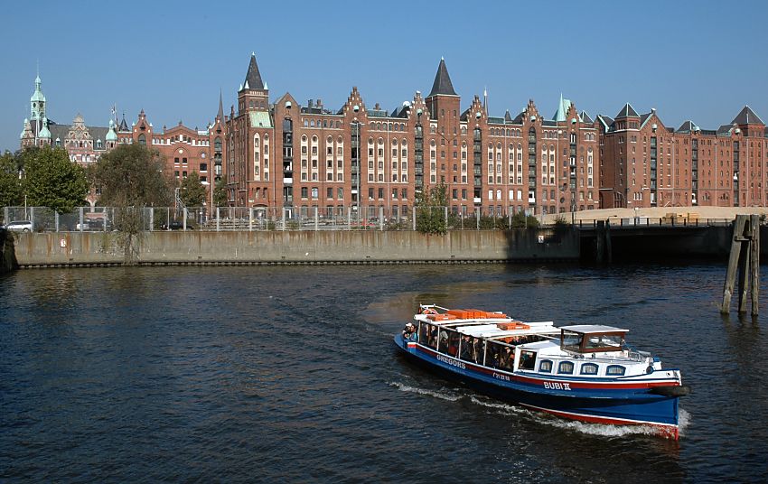 017_18947 Panorama der Hamburger Speicherstadt mit seiner Backsteinarchitektur und neogotischen Giebel und Trmen; lks. das sogen. Speicherstadtrathaus. Die Barkasse ist auf ihrer Fahrt Richtung Magdeburger Hafen und zur Elbe - rechts sind hlzerne Duckdalben zu erkennen, an denen frher Schiffe fest gemacht haben.  www.christoph-bellin.de