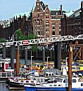 017_18959 Blick auf den Hamburger Binnenhafen; Barkassen liegen auf dem Wasser an den Dalben - im Hintergrund der Kehrwiedersteg, der zur Speicherstadt fhrt. Die kupfergedeckten Dcher der Speicherstadt schimmern in der Sonne. 