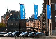 017_18987 drei Fahnenmaste mit den blauen Flaggen der Hafencity bei der Oberbaumbrücke - im Hintergrund die Speicherstadt im Sonnenlicht. In der hinteren Bildmitte ist die Spitze des Bürogebäudes des Hanseatic Trade Centers HTC zu erkennen.  ©www.christoph-bellin.de