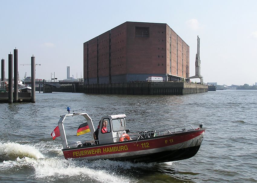 017_18996 ein schnelles Motorboot der Hamburger Feuerwehr verlsst seinen Standort am Kehrwieder und fhrt in rasanter Gleitfahrt auf die Elbe - im Heck des Boots wehen die Hamburg-Fahne und die "Nationale" / Deutschlandflagge im Fahrtwind. Im Hintergrund die massive Speicherarchitektur des Kaispeichers A. www.christoph-bellin.de