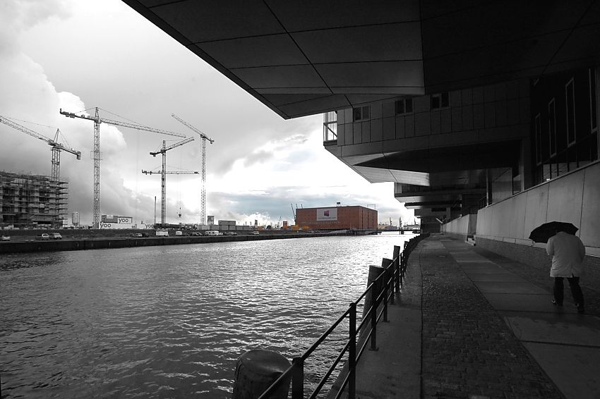 017_18999 Hamburger Regenwetter, ein Hamburg Tourist geht mit Regenschirm ientlang der Promenade am Sandtorkai / Sandtorhafen in der Hafencity - links die hohen Baukrne am Kaiserkai. Im Hintergrund der Kaispeicher A, der mit einem Glasaufbau versehen das neue Wahrzeichen Hamburgs, die Elbphilharmonie, werden soll.  www.christoph-bellin.de