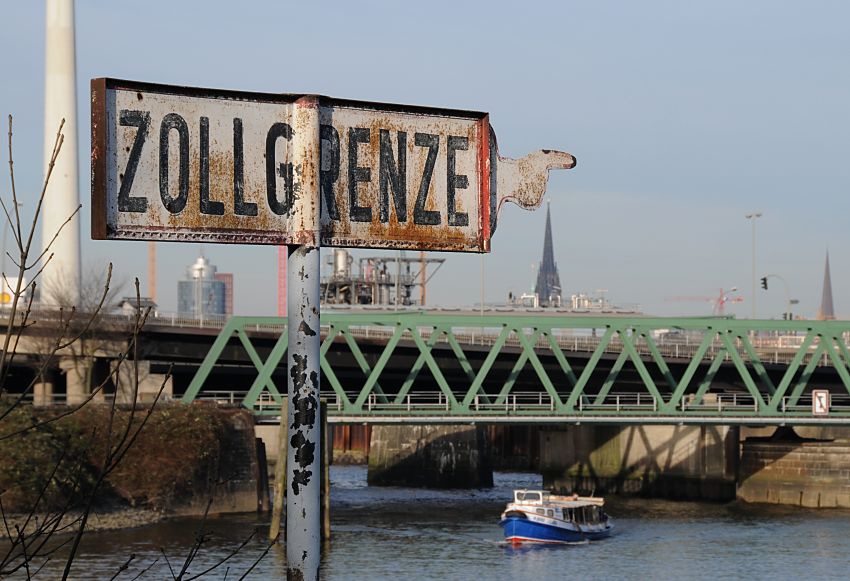 017_19011 ein altes Metallschild zeigt mit einer Hand und ausgestrecktem Finger den Verlauf der Zollgrenze - hamburg fotos : eine Barkasse der Hamburger Hafenrundfahrt kommt aus dem Reiherstieg und fhrt Richtung Ellerholzschleuse / Ellerholzhafen oder Kuhwrder Hafen. Im Hintergrund einige Trme von Hamburg: links das Brogebude des Hanseatic Trade Centers am Kehrwieder; unter der Hand die Turmruine der St. Nikolaikirche und rechts die St. Petrikirche.  www.christoph-bellin.de