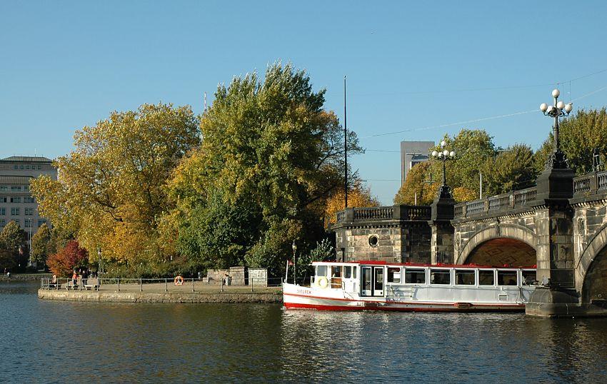 16_03821  Ein Alsterschiff passiert gerade die Lombardsbrücken - oben am Brückengeländer die typischen Kandelaber dieser historischen Hamburger Brücke, die zwischen Binnenalster und Aussenalster liegt. Links im Hintergrund der "Neue Jungfernstieg", rechts im Hintergrund das hohe Gebäude des Hotels am Dammtor.  ©www.christoph-bellin.de