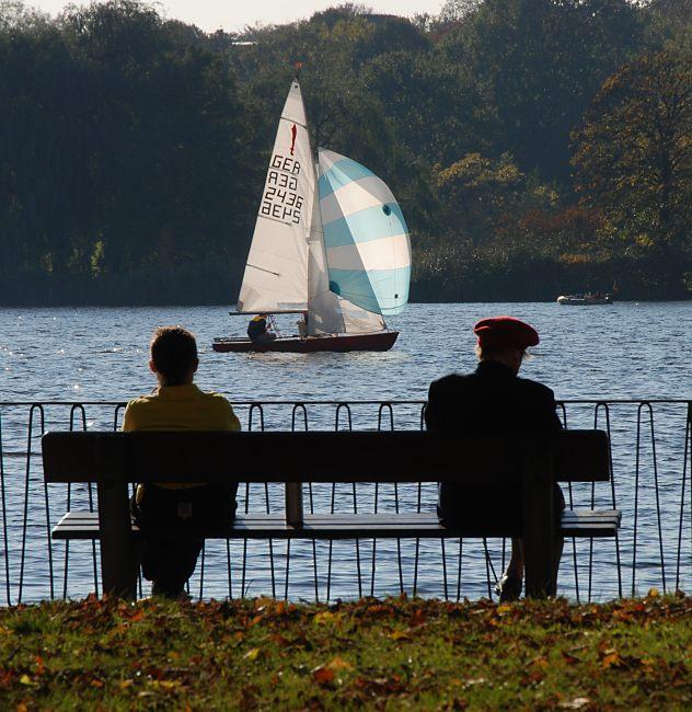 16_03822  Eine Parkbank am Alsterufer in Hamburg Uhlenhorst - zwei Besucher der Alster - Grnanlage  sitzen in der Sonne und beobachten ein Segelboot, dass unter vollen Segeln ber die Aussenalster Richtung Krugkoppelbrcke fhrt - im Vordergrund Herbstlaub auf der Wiese.  www.christoph-bellin.de