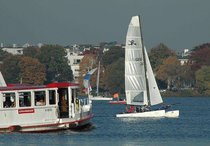 16_03824 ein Alsterschiff der Alster-Rundfahrt fhrt mit den Gsten an Bord ber die Alster Richtung Anleger Rabenstrasse - ein weisser Katamaran kreuzt das Fahrgastschiff. Im Hintergrund Herbstbume mit ihren roten, gelben und goldenen Blttern.   www.christoph-bellin.de