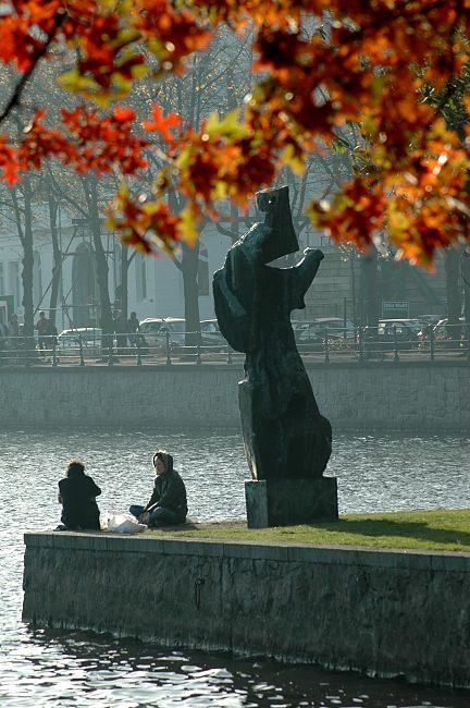 16_03816 zwei HamburgerInnen sitzen in der herbstlichen Abendsonne auf der Mauer am Ufer der Binnenalster; die Skulptur "Die Windsbraut" von Hans-Martin Ruwoldt wirft einen langen Schatten - davor das rotbraune und goldene Herbstlaub von einem Ahorn.  www.christoph-bellin.de