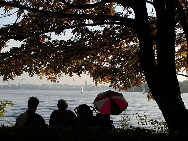 16_03816 eine Familie sitzt am Ufer der Aussenalster unter einem herbstlich gefrbten Baum und blickt auf die wenigen Segelboote, die noch auf dem Wasser sind. An der Kinderkarre / Kinderwagen ist der Sonneschirm zum Schutz aufgespannt. Im Hintergrund das Panorama von Hamburg im Abenddunst mit Kirchtrmen und Baukrnen.  www.christoph-bellin.de