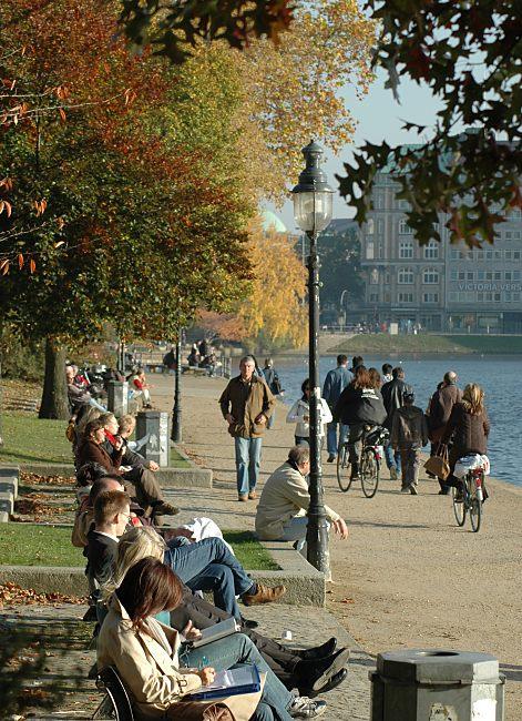 16_03818 Mittagsstunde in der Hamburger Innenstadt; die Hamburger sitzen in auf Parkbnken in der Sonne an der Binnenalster bei der Lombardsbrcke - Fahrradfahrer fahren auf dem Fussweg Richtung Aussenalster. Im Hintergrund Gebude am Ballindamm.   www.christoph-bellin.de