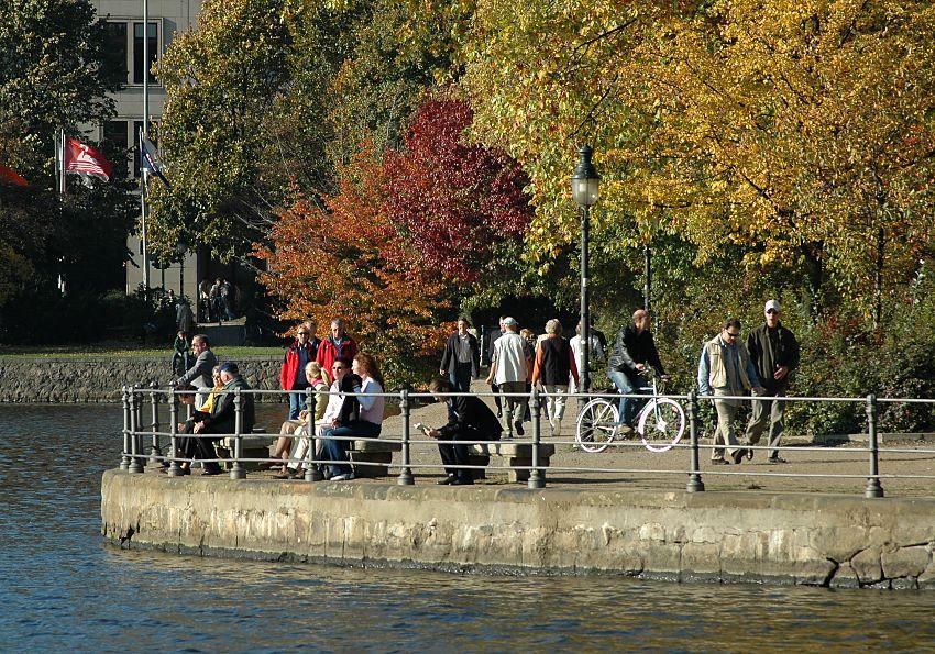 16_03819  prchtig gefrbt Herbstbume und Strucher in der Sonne an Hamburgs Binnenalster - die Sitzbnke in der Sonne sind von von Hamburgern besetzt, die die warmen Sonnenstrahlen der Herbstsonne geniessen - Spaziergnger und Spaziergngerinnen schlendern auf der Promenade an den Lombardsbrckenr; ein Fahrradfahrer fhrt Richtung Aussenalster.    www.christoph-bellin.de