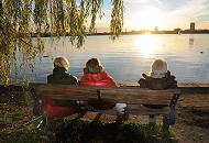 426_1369  Die Herbstsonne steht niedrig ber der Aussenalster, ein vereinzeltes Segelboot fhrt an diesem spten Herbstnachmittag auf der Alster. Unter einer Weide, deren Bltter schon teilweise eine Herbstfrbung angenommen haben, ruhen sich drei Spaziergngerinnen auf einer Holzbank aus und blicken ber das Wasser des Sees in die Sonne. 