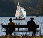 16_03822  Eine Parkbank am Alsterufer in Hamburg Uhlenhorst - zwei Besucher der Alster - Grnanlage  sitzen in der Sonne und beobachten ein Segelboot, dass unter vollen Segeln ber die Aussenalster Richtung Krugkoppelbrcke fhrt - im Vordergrund Herbstlaub auf der Wiese.  www.christoph-bellin.de