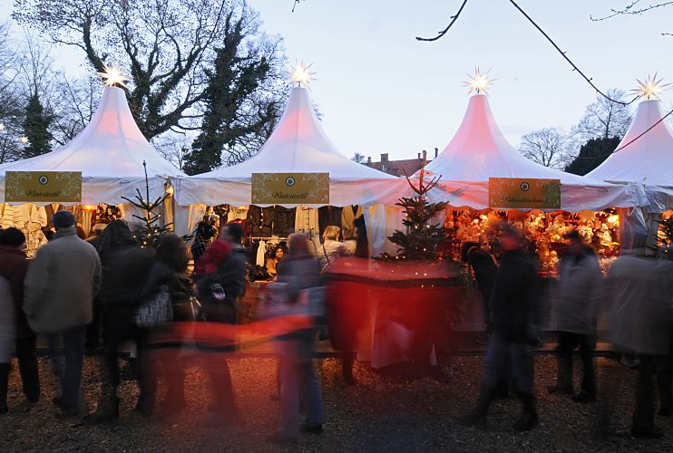 Foto von Marktständen auf dem Bergedorfer Weinachtmarkt am Schloss  Die Besucher und Besucherinnen schlendern am frühen Abend über den Bergedorfer Weihnachtsmarkt. Die Marktstände sind festlich beleuchtet und mit Tannenbäumen und beleuchteten Sternen geschmückt.