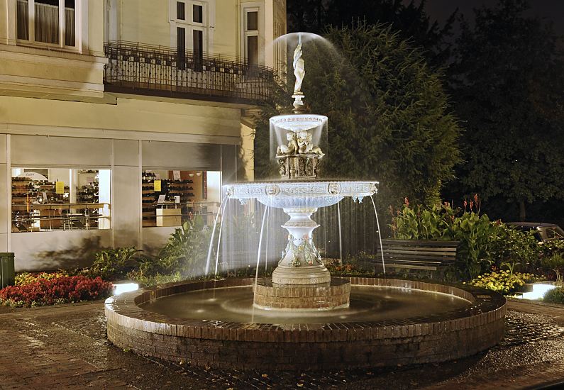 Fotografie vom beleuchteten Springbrunnen / Sievers - Brunnen am Kaiser Wilhelm Platz.  Abends wird der Springbrunnen auf dem Kaiser Wilhelm Platz beleuchtet. Die zwei Wasserbecken aus Gusseisen und die einzelnen Dekorelemente der Wasseranlage sind farblich gefasst.