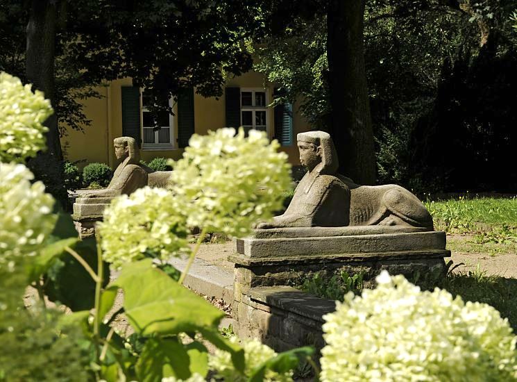 Bild vom Rathauspark in Bergedorf - Sphinxen und Hortensienblten.  Hinter den weissen Blten von Hortensien zwei Sphinx - Skulpturen im Garten des Bergedorfer Rathauses; zwischen den Sphingen fhren in der Gartenanlage Stufen zum  Rathausgebude. Im Hintergrund ist das Bergedorfer Standesamt zu erkennen - die Fenster haben Fensterladen aus Holz.