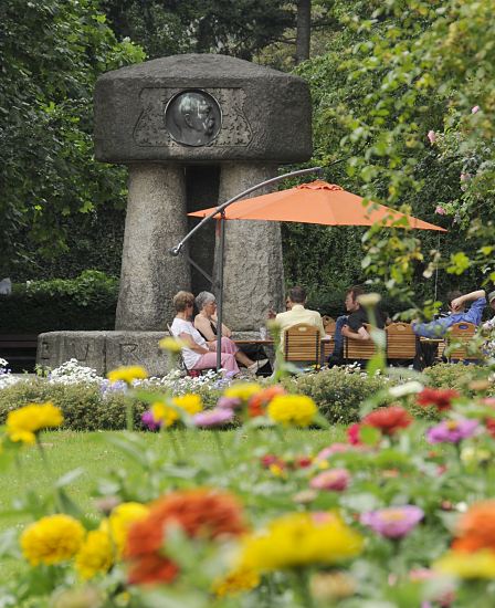 Foto Bismarck Denkmal Bergedorfer Schlosspark  Das Bergedorfer Bismarck Denkmal wurde 1906 eingeweiht, den Entwurf fr die monumentale Erinnerungssttte lieferte Karl Garbers. Das Bismarkdenkmal wurde 1906 eingeweiht -  erst spter wurde es in den Bergedorfer Schlosspark versetzt. Im Vordergrund blhen bunte Blumen - Parkbesucher sitzen unter einem Sonnenschirm in einem Caf neben dem Granitdenkmal an dessen Stirnseite die Kupferplakette mit dem Kopf Bismarcks.