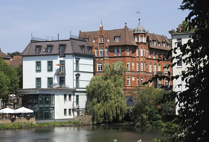  Blick zum Wehr an der alten Holstenstrasse - am Wasserdurchlass der Bille in den Bergedorfer Hafen steht eine Weide deren ste tief ber dem Wasser hngen. Im Hintergrund ein roter Klinkerbau mit Erker und Giebelturm, lks. ein Grnderzeitgebude an dessen Wasserseite Gste eines Bistros auf einer Terrasse in der Sonne sitzen knnen. Sommerbilder aus Hamburg Bergedorf - Bergedorfer Hafen, Serrahn in der Sonne.