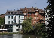 17_21563 Blick zum Wehr an der alten Holstenstrasse - am Wasserdurchlass der Bille in den Bergedorfer Hafen steht eine Weide deren ste tief ber dem Wasser hngen. Im Hintergrund ein roter Klinkerbau mit Erker und Giebelturm, lks. ein Grnderzeitgebude an dessen Wasserseite Gste eines Bistros auf einer Terrasse in der Sonne sitzen knnen. www.hamburg-fotograf.com