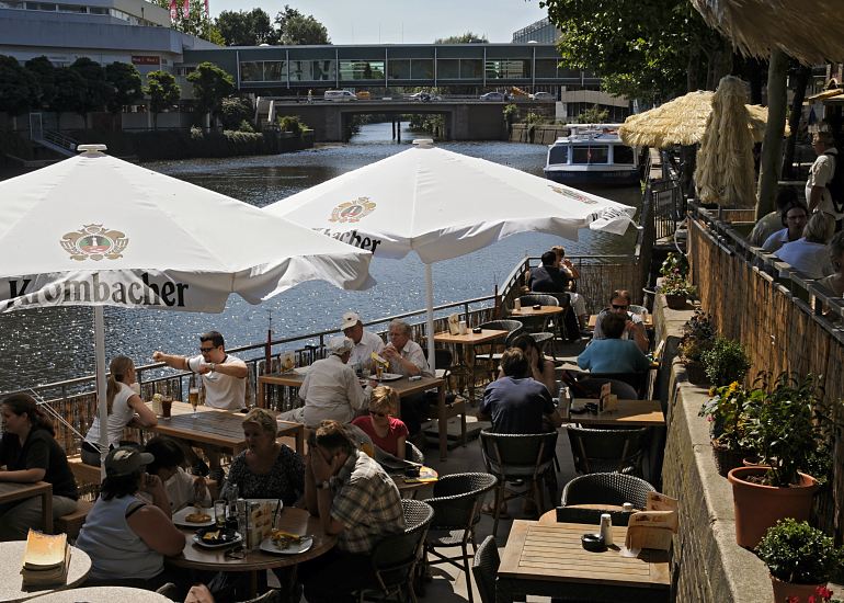 Blick ber den Bergedorfer Hafen - ein Fahrgastschiff der Bergedorfer Schifffahrtslinie liegt am Kais vom Serrahn. Auf der Aussenterrasse eines Bistros am Hafen sitzen die Gste in der Sonne oder Sonnenschirmen. Im Hintergrund ist unter der Brcke die Zufahrt zum Schleusengraben zu erkennen.  Sommer in Hamburg, Aussenterrasse mit Sonnenschirmen am Bergedorfer Hafen.