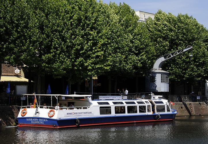 Das Ausflugsschiff SERRAHN DEERN liegt an der Kaimauer des Bergedorfer Hafens - Fahrgste sitzen im offenen Heck der Barkasse und warten auf den Beginn der Rundfahrt durch die Vierlnder Wasserwege. Dahinter der elektrische Drehkran, der 1901 bei der Fabrik Nagel & Kaemp in Hamburg Winterhude gefertigt wurde. Der historische Hafenkran steht unter Denkmalschutz und ist Zeugnis aus der Zeit als im Bergedorfer Hafen Lastschiffe be- und entladen wurden. Fahrgastschiff SERRAHN DEERN an der Anlegestelle Serrahn, historischer Drehkran.