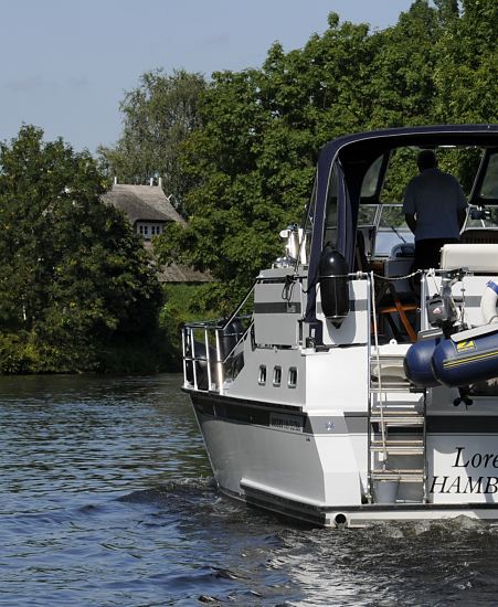 Hamburg Fotografie - Motorboot auf der Doveelbe  Das weisse Motorboot fhrt in der Flussmitte der Dove- Elbe; der Freizeitkapitn steht am Steuerstand unter der Persenning als Schutz gegen die Sonne. Hinter dem Deich ist das Strohdach eines Wohnhauses zu erkennen.