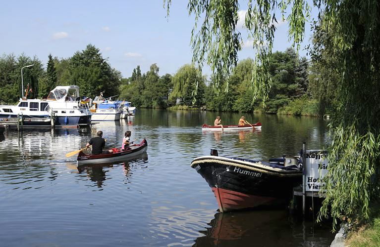 Zwei Kanus fahren bei strahlendem Hamburger Sommerwetter auf der Dove-Elbe - lks. linken liegen Motoryachten am Bootssteg eines Vierlnder Yachthafens. Im Vordergrund am Steg des Hotels Vierlandentor hat das Tuckerboot HUMMEL festgemacht - ein dicker umlaufenden Tampen / Seil dient als Scheuerschutz. Die Tuckerboote waren offene Arbeitsbarkassen im Hamburger Hafen; sie brachten als Zubringerboote u.a. auch Hafenarbeiter an ihren Arbeitsplatz.  Hamburg Fotograf - Motive der Flsse Hamburgs - Kanus und Yachthafen an der Dove-Elbe