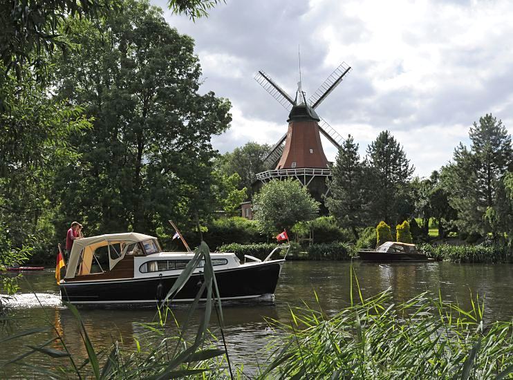 Die Dove-Elbe ist ein beliebtes Hamburger Gewsser fr Freizeitkapitne. Ein Motorboot fhrt flussabwrts, am Bug fhrt es die Hamburg-Fahne und am Heck die Deutschlandflagge. Die kleine Motoryacht passiert gerade die historische Reitbrooker Windmhle. Die Kornmhle wurde  ursprnglich 1773 errichtet und nach einem Brand 1870 neu erbaut. Am Ufer im Vordergrund wchst dichtes Schilf - auf der anderen Uferseite liegt am Bootsanleger ein kleines Sportboot.  Fotos Sportbootrevier Hamburger Doveelbe - Reitbrooker Windmhle, Motorboot 