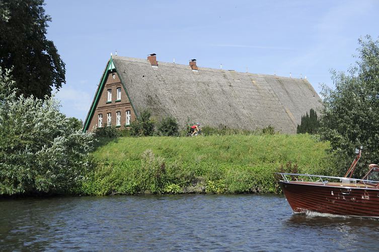 Das Dach eines reetgedeckten Bauernhauses ragt hinter dem Deich der Doveelbe hervor. Die Deichstrasse ist in Hamburg eine sehr beliebte Strecke fr die bungsfahrten von Radrennfahrern. Eine Motoryacht fhrt flussabwrts auf der Dove-Elbe  Hamburg Fotografie - Sommer in Bergedorf - Strohdachhaus am Deich, Motorboot