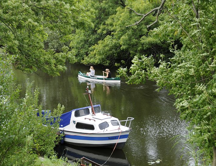 Blick auf die Dove-Elbe bei Hamburg Reitbrook; das Ufer des Flusses ist dicht mit hohen Bäumen bewachsen - die Äste hängen dicht über dem Wasser. Ein Kanu fährt auf dem ruhigen Wasser der Doeveelbe, im Vordergrund ist ein kleines Motor-Sportboot am Bootssteg vertäut.  Grünes Hamburg Bergedorf - Bäume am Ufer der Dove Elbe - Kanufahrt