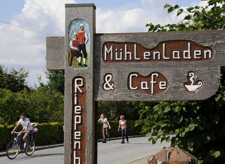 Handgeschnitztes Hinweisschild zum Mhlenladen und Cafe bei der Riepenburger Mhle. Auf der Strassen fahren Inlineskater und ein Fahrrad. Die Strassen durch die Drfer der Vier- und Marschlande werden am Wochenende hufig von den Hamburger Ausflglern fr dieses sportliche Freizeitvergngen genutzt  Hamburg Bilder, Bezirk Bergedorf - Schild Mhlenladen & Cafe Riepenburger Mhle.