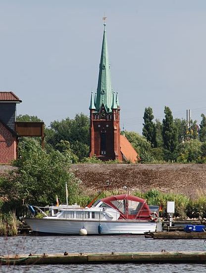 Fotografie Hamburg - Kirchturm St. Nikolai   Der Kirchturm der St. Nikolaikirche in Hamburg Moorfleet wurde 1885 errrichtet. Im Vordergrund ein Sport- boot mit herunter geklapptem Verdeck und Bootsstege im Holzhafen; dahinter ist der Deich zu erkennen