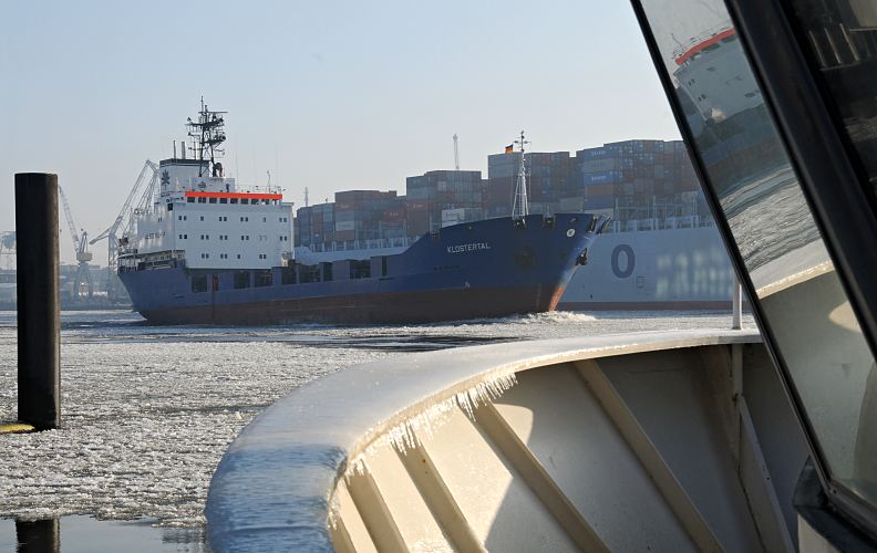 Fotos vom Hamburger Winter auf der Elbe - Containerfeeder KLOSTERTAL. 004_22733 Der niederländische Containerfeeder Klostertal fährt durch den Eisgang auf der Elbe flussabwärts - das Schiff spiegelt sich in der Scheibe einer Hafenfähre. Hinter dem Frachtschiff läuft ein hoch beladener Containerfrachter in den Hamburger Hafen ein.