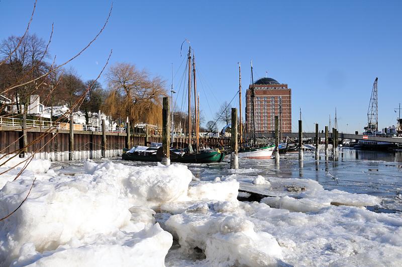 06_5723 Hoch aufgetrmt liegen die Eisschollen am Ufer des im Museumshafens im Hamburg Oevelgoenne. Einige der historischen Schiffe berwintern auf dem Wasser, andere lagern an einem eissicheren Winterplatz. Hinter dem Steg zum Fhranleger der Hafenfhre liegt das Gebude des ehemaligen Khlhauses in Neumhlen. Bilder vom Hamburger Winter - vereister Museumshafen velgnne; historische Segelschiffe. 