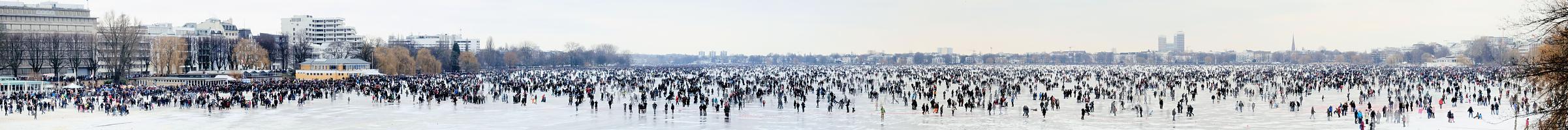 2021_31 Panoramblick auf die zugefrorene Aussenalster - ab einer Strke von 20cm darf das Eis des grossen Hamburger Sees offiziell betreten werden. Es findet das sogenannte Alstereisvergngen statt; Buden mit Glhwein und Wrstchen stehen am Ufer der Alster. Hunderttausende HamburgerInnen und Hamburg-Touristen gehen auf dem zugefrorenen See spazieren, laufen Schlittschuh oder spielen Eishockey.  