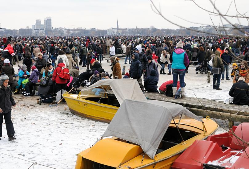 2058 Dicht an dicht gehen die Menschen auf der zugefrorenen Alster; Kinder spielen auf dem Eis. Am Holzsteg liegen einige Boote einer Bootsvermietung - die kleinen Schiffe sind im dicken Eis eingefroren. 