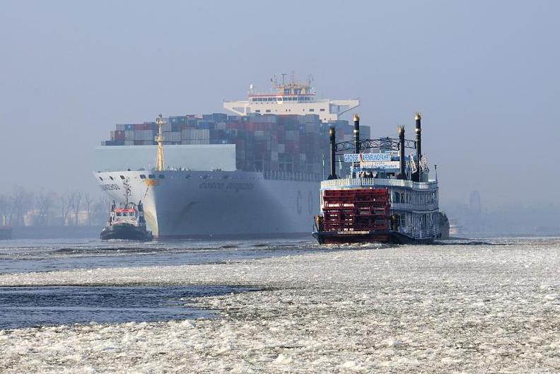 2988 Der Containerfrachter COSCO GUANGZHOU luft in den Hamburger Hafen ein - ein Schlepper dirigiert den Containerriesen im Fahrwasser der Elbe. Ein Raddampfer der Groen Hafenrundfahrt fhrt durch das Elbeis auf seiner Tour durch den vereisten Hafen Hamburgs.