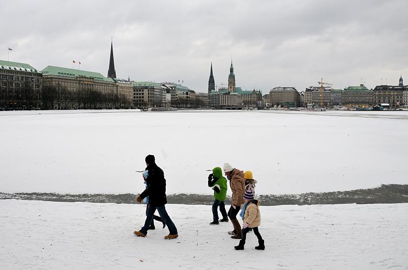 31_4983 Eine Familie geht warm angezogen an einem grauen Wintertag an der Binnenalster spazieren. Im Hintergrund der Rathausturm und die Kirchtrme von St. Petri, St. Nikolai und der St. Michaeliskirche. Hamburgbilder vom Winter - Familienspaziergang an der Binnenalster bei Eis und Schnee.