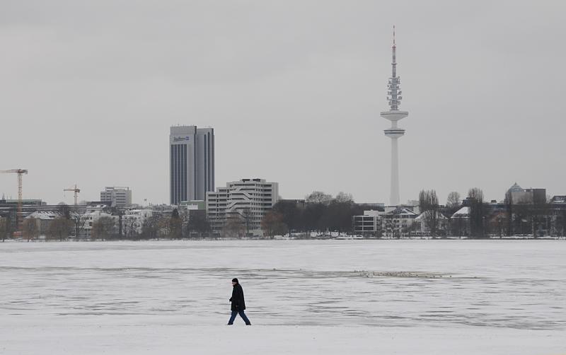 34_4981 An einem grauen Wintertag geht ein einsamer Spaziergnger am Ufer der zugefrorenen Alster spazieren. Am anderen Ufer des 164 ha groen Hamburger Binnensees ragt der Fernsehturm und das Radisson Hotel in den Winterhimmel. Aufnahmen von Hamburger im Winter - grauer Wintertag am Alsterufer.