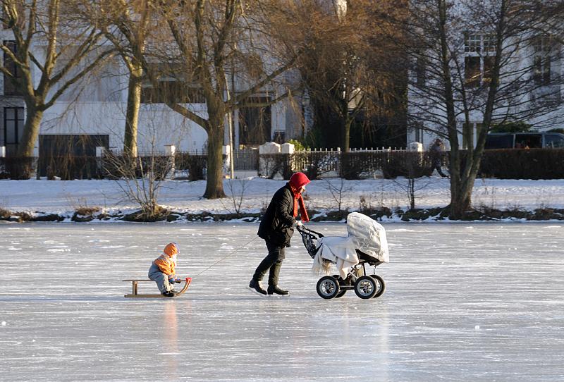 38_5468 Eine Mutter fhrt Schlittschuh auf dem Eis der Alster - sie schiebt dabei einen Kinderwagen und zieht einen Schlitten, auf dem ein Kind sitzt und die Fahrt in der Sonne geniet.  Hamburger Winterbilder - Schlittschuhlaufen mit Kinderwagen und Schlitten auf der Alster.