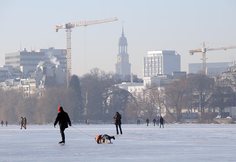 40_5509 Spaziergnger genieen das schne Winterwetter und gehen in der Wintersonne auf der zugefrorenen Alster spazieren. Eine Mutter luft Schlittschuh und zieht dabei ihr Kind auf einem Schlitten. Im Hintergrund der Turm der St. Michaeliskirche.  Hamburger Wintermotive vom Winter 2009 / 2010 - die Hamburger Auenalster ist zugefroren.