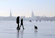 43_5501 Hamburger nutzen das schne Winterwetter und gehen mit ihrem Hund ber das blanke Eis der zugefrorenen Alster. Im Hintergrund die Kennedybrcke und die Trm der St. Petrikirche und St. Nikolaikirche sowie der Turm des Hamburger Rathauses. Hamburgbilder im Winter - Spaziergnger mit Hund auf der zugefrorenen Alster. 