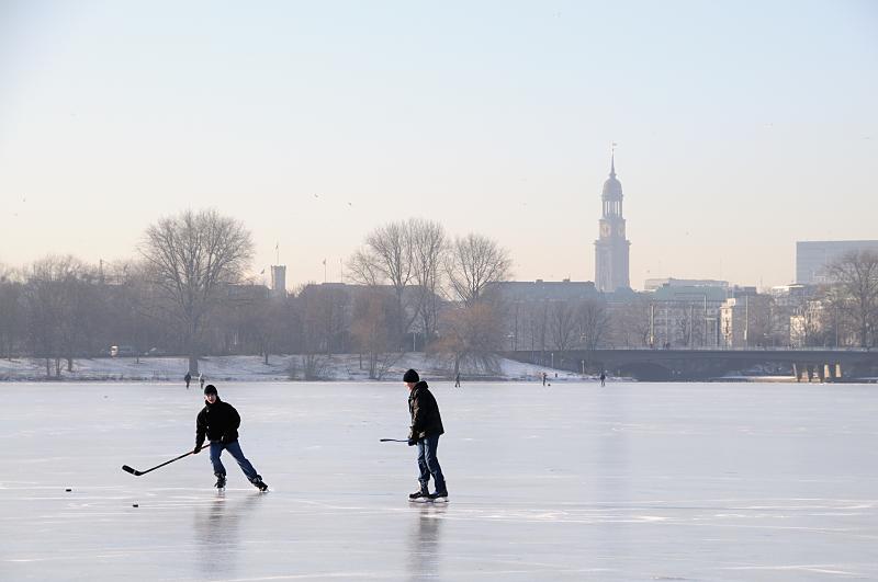 44_5574 Hamburger Jugendliche spielen Eishockey auf der zugefrorenen Alster - hinter der Kennedybrücke sind die Häuser am Jungfernstieg zu erkennen und der Kirchturm der St. Michaeliskirche. Hamburger Jahreszeiten, die Hansestadt im Winter - Eishockeyspieler auf der Außenalster. 