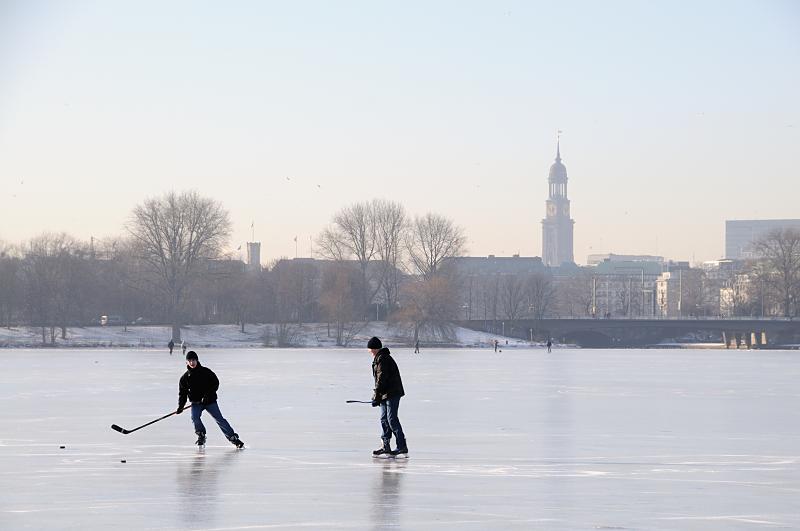 44_5574 Hamburger Jugendliche spielen Eishockey auf der zugefrorenen Alster - hinter der Kennedybrcke sind die Huser am Jungfernstieg zu erkennen und der Kirchturm der St. Michaeliskirche. Hamburger Jahreszeiten, die Hansestadt im Winter - Eishockeyspieler auf der Auenalster. 