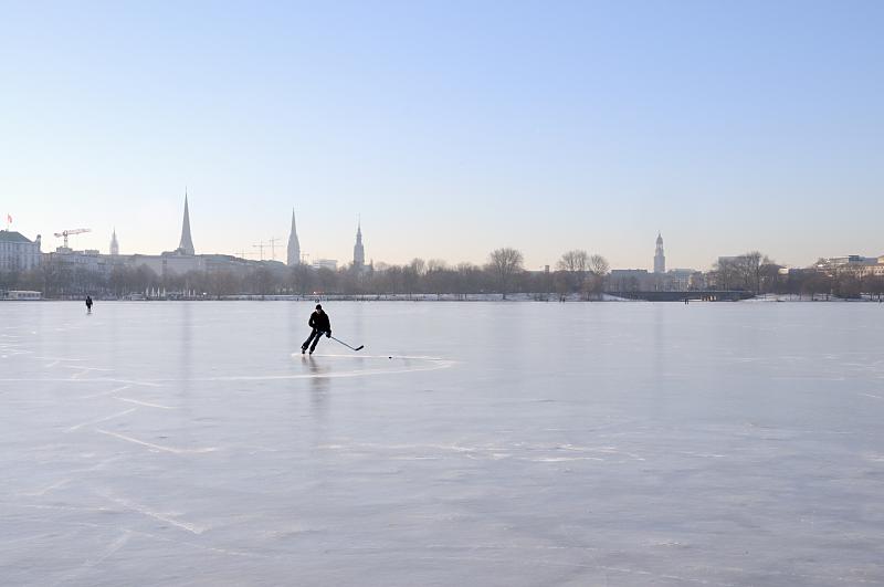 45_5558 Ein Eishockeyspieler dreht seine Kurven auf dem Eis der Alster bei frostigem Winterwetter. Im Hintergrund das Panorama von Hamburg mit den Trmen der Hansestadt.  Die Hansestadt Hamburg im Winter - Panorama der Stadt, Eishockeyspieler auf der Alster.