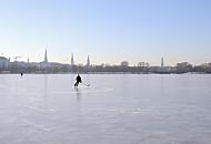 45_5558 Ein Eishockeyspieler dreht seine Kurven auf dem Eis der Alster bei frostigem Winterwetter. Im Hintergrund das Panorama von Hamburg mit den Trmen der Hansestadt.  Die Hansestadt Hamburg im Winter - Panorama der Stadt, Eishockeyspieler auf der Alster.