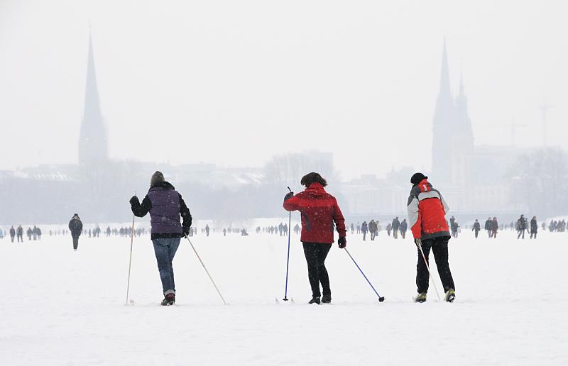 48_6109 Skilangluferinnen nutzen den hohen Schnee auf dem Alsterei, um eine Runde um den 164 ha groen Binnensee zu machen; die Rundstrecke am Alsterufer ist fr Jogger ca. 7,5 km lang. Im Hintergrund in den Schneewolken die Kirchtrme der St. Petri und Nikolaikirche sowie rechts der Rathausturm. Fotos vom Hamburger Winter - HamburgerInnen machen Skilanglauf auf der Alster.