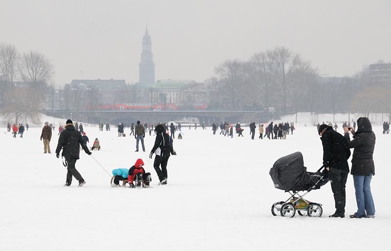 49_6117 Die Hamburgerinnen und Hamburger genieen mit ihren Kinder das Alstereisvergngen -sie ziehen ihre Sprsslinge auf Schlitten ber das Eis oder schieben ihren Kinderwagen ber die Alster. Im Hintergrund der Michelturm - ber die Lombardsbrcke fhrt ein Zug. Winterbilder aus Hamburg - Winterspass im Schnee auf der zugefrorenen Alster.