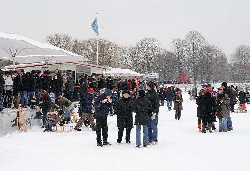 50_6130 Auf dem Alsterponton, an dem normalerweise die Alsterschiffe anlegen, stehen die Hamburger Alsterbesucher Schlange, um an einem der Verkaufsstnde heien Glhwein oder eine Bratwurst zu kaufen. Andere stehen im Schneetreiben auf dem Eis der Alster und genieen die Hamburger Winteratmosphre. Winterfotos aus Hamburg - Alstereisvergngen bei Glhwein und Bratwurst im Schneegestber.