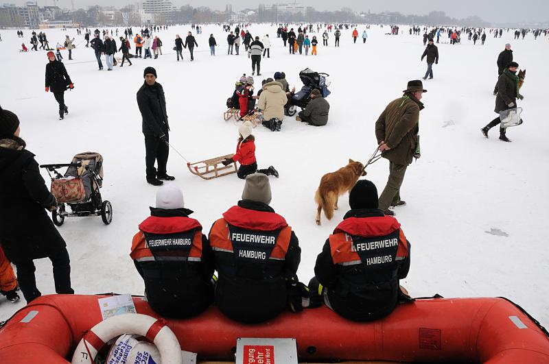 51_6156 Am Rande des Alstereisvergngens sitzen Rettungskrfte der Feuerwehr Hamburger auf ihrem Schlauchboot und beobachten das Treiben auf dem Eis. Falls jemand in das Eis einbrechen sollte, wrden sich die Feuerwehrmnner mit Hilfe des Schlauchboots an die Einbruchsstelle heranschieben und die verunglckte Person in Sicherheit bringen. Hamburg im Winter - Rettungskrfte der Hamburger Feuerwehr am Rand der zugefrorenen Alster. 