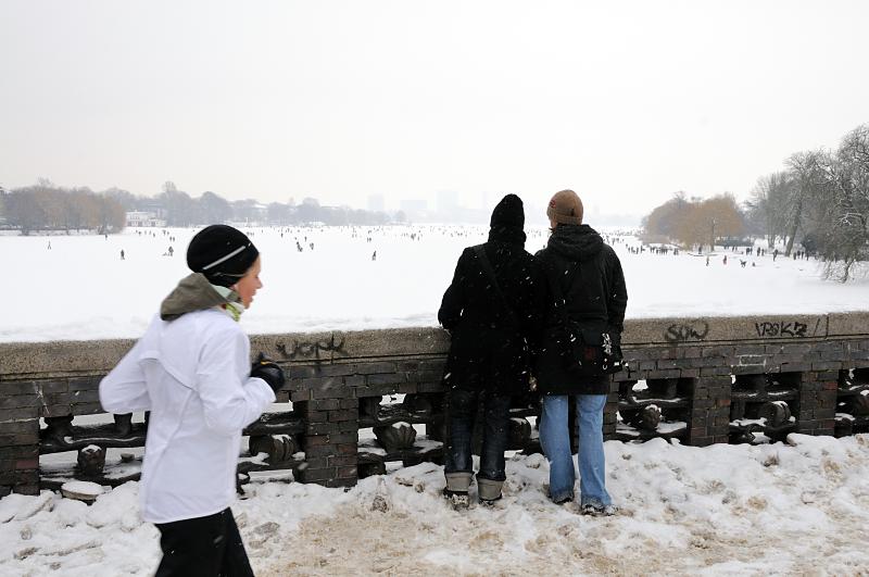 52_6076 Von der Krugkoppelbrcke hat man einen weiten Blick ber die weie, zugefrorene Alster - die Hamburger nutzen das Wintereis, um quer ber die Alster in die Hamburger Innenstadt zu spazieren. Andere joggen an Land durch den Schnee die 7,5 km um Alster herum. Aufnahmen von Hamburg im Winter - Blick von der Krugkoppelbrcke auf die Alster, Joggerin. 